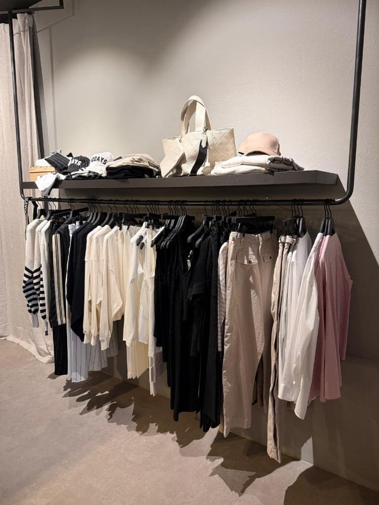 Kleiderstange mit Blusen, Strick und Hosen in Schwarz, Creme und Beige, darüber Taschen und Caps